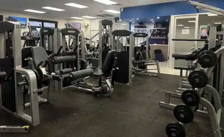 Pin-loaded machines and a dumbbell rack on the strength floor, with the blue Vitality reception wall behind