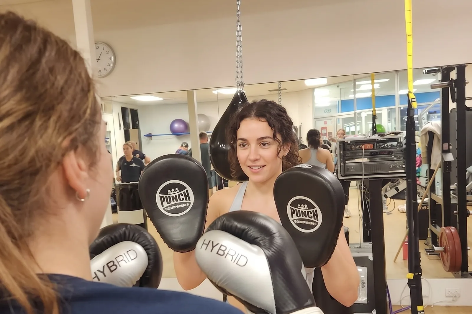A coach holding pads for a member during a boxing session, both mid-round