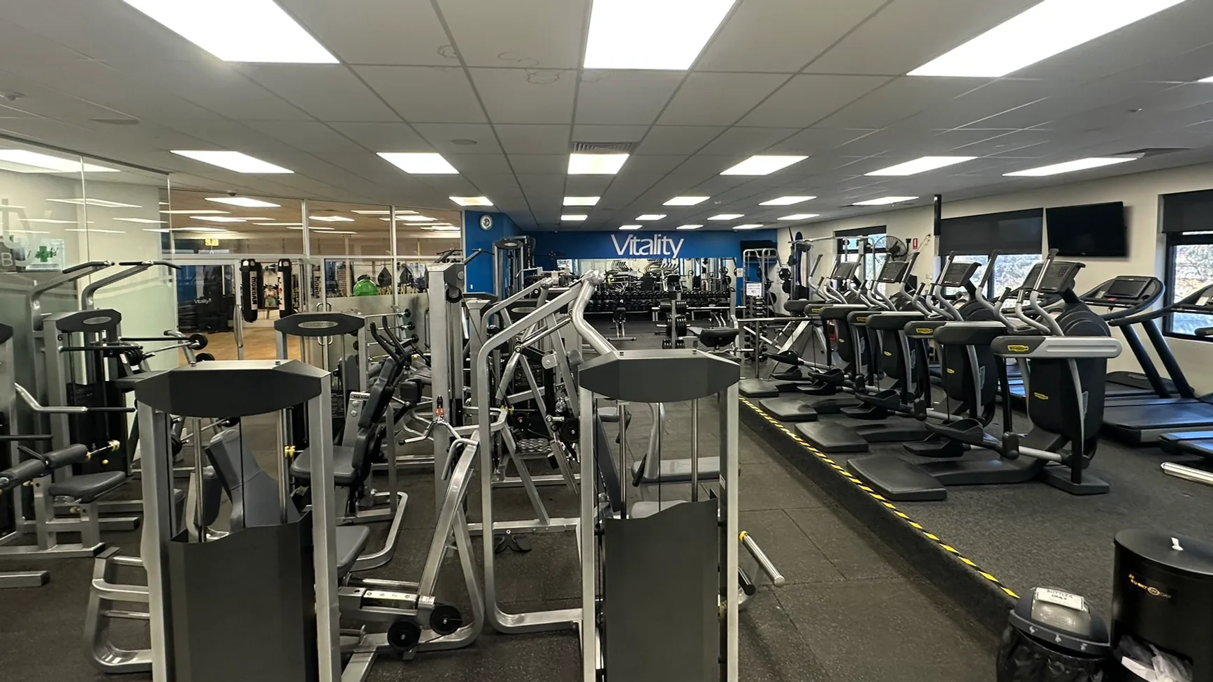 The training floor at the Berri club, shot wide from the entrance: strength machines in the foreground, the cardio row along the windows and the Vitality wall at the far end
